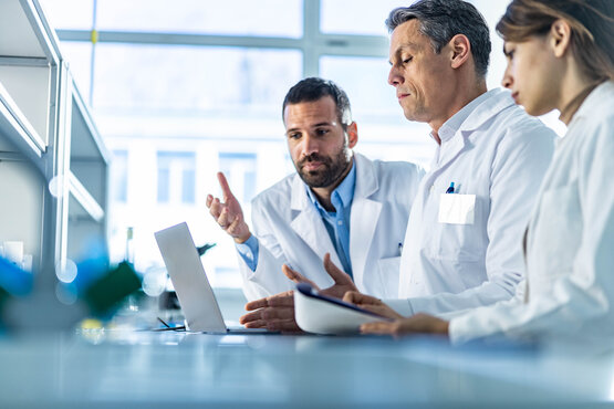 Zwei Ärzte und eine Ärztin stehen vor einem Laptop und halten Dokumente in den Händen
