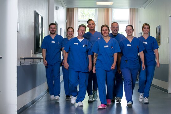 Acht Personen in blauen medizinischen Uniformen gehen gemeinsam einen hellen Krankenhausflur entlang