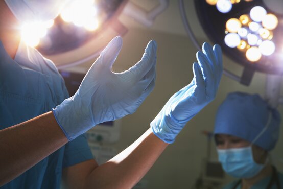 Zwei Personen in blauer OP-Kleidung, eine hebt behandschuhte Hände vor OP-Licht, eine mit Maske und Haube im Operationssaal