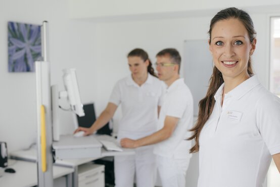 Drei Personen in weißer Kleidung, eine Frau mit Pferdeschwanz im Vordergrund, zwei am Schreibtisch mit Monitor im hellen, modernen Praxisraum