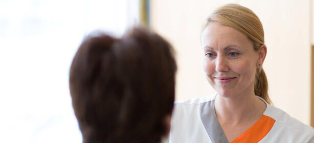 Freundliche Pflegekraft mit blondem Haar im Gespräch mit Patientin, beide in heller Umgebung, empathische Pflege