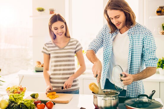 Zwei junge Erwachsene kochen gemeinsam in heller, moderner Küche; Frau schneidet Gemüse, Mann kocht Nudeln