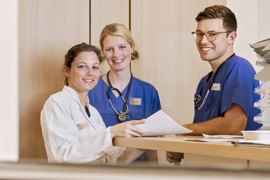 Drei medizinische Fachkräfte lächeln am Schreibtisch mit Dokumenten und Aktenordnern in der Klinik