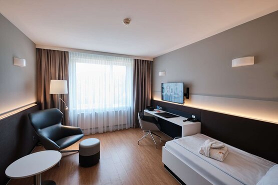 Einzelzimmer mit Bett, Schreibtisch, Sessel, Fernseher und Fenster
