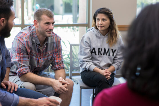 Erwachsene sitzen in Stuhlkreis in hellem Raum, Mann im karierten Hemd und Frau im „ARMY“-Hoodie hören aufmerksam zu, Gruppentherapie
