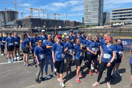 Große Gruppe in blauen T-Shirts mit Startnummern bei Laufevent am Wasser, Baukräne und moderne Gebäude im Hintergrund