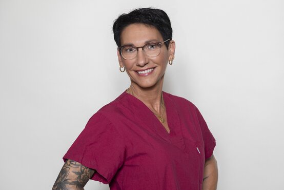 Pflegekraft mit kurzen dunklen Haaren, Brille und Tattoo im dunkelroten Kasack lächelt vor hellem Hintergrund in die Kamera