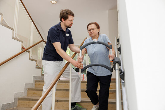 Ältere Frau trainiert mit Physiotherapeut an einer Treppe mit Handläufen in der Frührehabilitation.