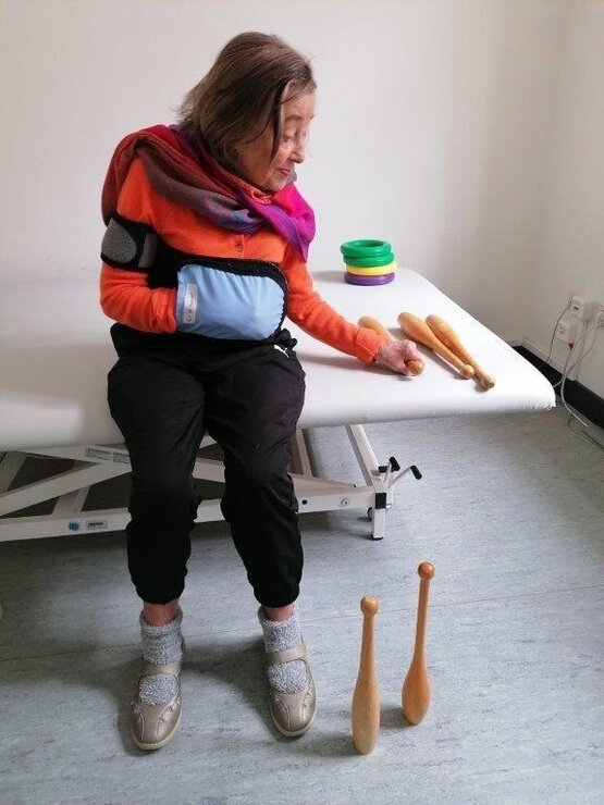 Frau mit Armschlinge trainiert auf Therapieliege mit Holzkeulen und bunten Ringen in der Frührehabilitation.