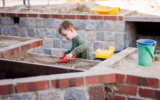 Kind spielt auf erhöhtem Sandkasten mit einer Schaufel auf Klinikspielplatz, Ansicht von vorne