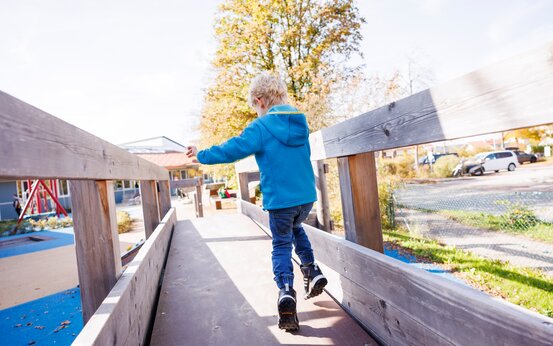 Blondes Kind in blauer Jacke läuft auf breiter Brücke mit Holzgeländer auf Klinikspielplatz