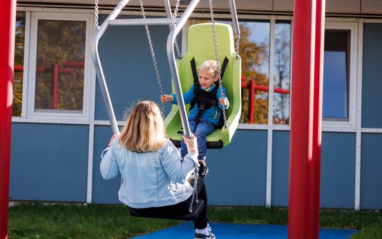 Mutter und Kind schaukeln auf behindertengerechter Schaukel auf Klinikspielplatz