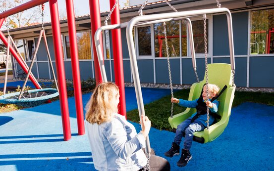 Mutter und Kind schaukeln auf behindertengerechter Schaukel auf Klinikspielplatz, Seitenansicht