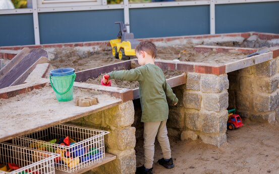 Kind spielt auf erhöhtem Sandkasten mit einer Schaufel auf Klinikspielplatz, Ansicht von hinten