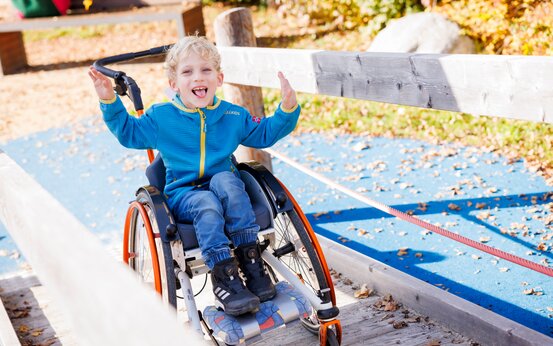 Junge mit blonden Locken im Rollstuhl auf barrierefreier Rampe auf Klinikspielplatz