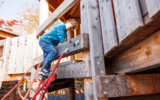 Junge mit blauer Jacke und Jeans klettert an einem roten Kletternetz auf Klinikspielplatz; umgeben von Holzbalken und Geländern