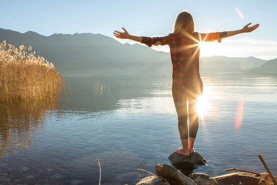 Person steht barfuß auf Stein am klaren See vor Bergkulisse, ausgebreitete Arme