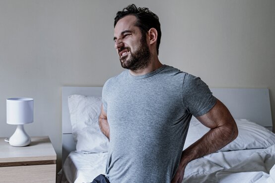 Mann sitzt auf einem Bett und hält sich schmerzhaft den Rücken