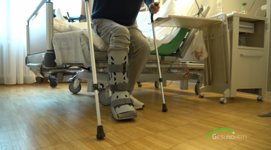 Patient mit Orthese am linken Bein steht nach Haglundferse-OP mit Unterarmgehstützen im Krankenhauszimmer auf
