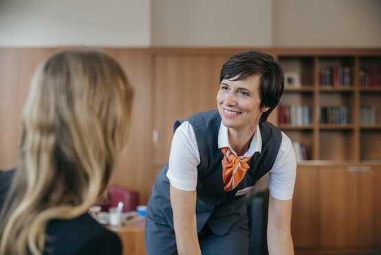 Freundliche Mitarbeiterin mit orangefarbenem Halstuch lächelt Person herzlich zu