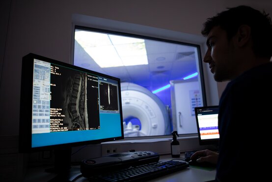 Radiologe am Arbeitsplatz analysiert MRT-Aufnahmen der Wirbelsäule, im Hintergrund MRT-Scanner mit blauer Beleuchtung.