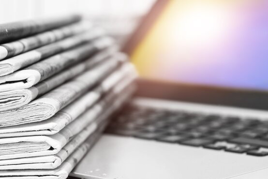 Stapel gefalteter Tageszeitungen in Schwarz-Weiß liegt auf der Tastatur eines Laptops mit beleuchtetem Bildschirm im Hintergrund