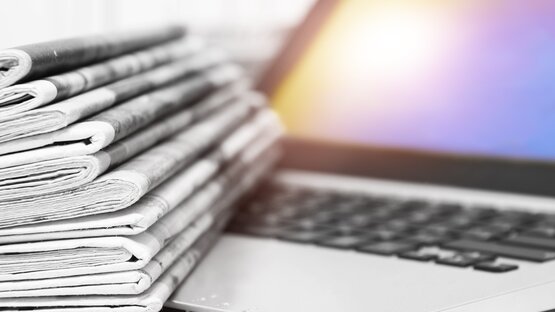 Stapel gefalteter Tageszeitungen in Schwarz-Weiß liegt auf der Tastatur eines Laptops mit beleuchtetem Bildschirm im Hintergrund