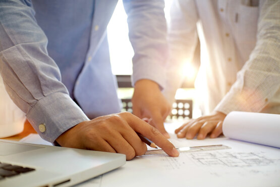 Zwei Personen in Hemden beugen sich über Tisch mit Bauplänen, eine Hand zeigt auf Plan, Laptop am Rand, Sonnenlicht im Hintergrund