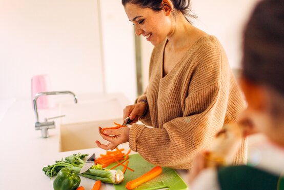 Frau schält lächelnd Karotte in heller Küche, frisches Gemüse auf Arbeitsplatte