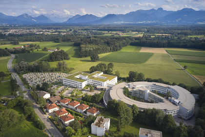 Luftaufnahme der modernen Klinik mit ringförmigem Gebäude, grüner Umgebung und Alpen im Hintergrund