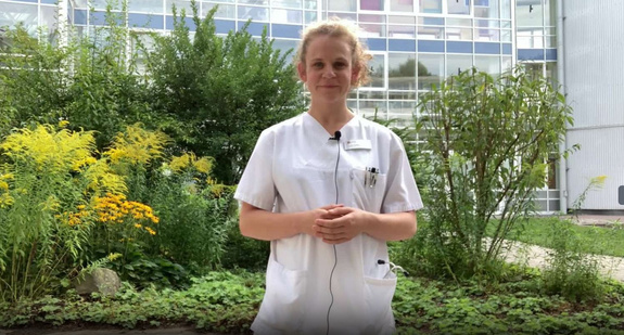 Lydia Patzak. Junge Frau im weißen Kittel mit Mikrofon und Ausweis steht lächelnd vor moderner Klinik
