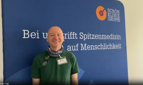 Markus Tillmann. Lächelnder Mitarbeiter der Schön Klinik im grünen Poloshirt vor blauem Hintergrund mit Kliniklogo