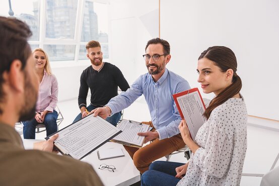 Fünf Erwachsene sitzen im Halbkreis im hellen Büro, halten Klemmbretter und lächeln sich an