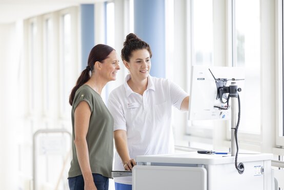 Zwei Frauen schauen gemeinsam auf Computer, medizinisches Personal erklärt Patientin digitale Abläufe
