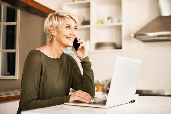 Frau mit kurzen blonden Haaren telefoniert lächelnd am Smartphone und arbeitet am Laptop