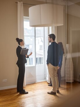 Wohnungsbesichtigung: Immobilienmaklerin erklärt jungem Paar helle, leere Wohnung mit Parkett und großen Fenstern