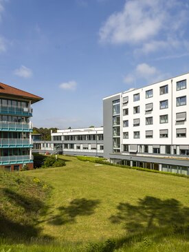 Moderne Klinikgebäude mit großen Fenstern und Balkonen, umgeben von gepflegter grüner Rasenfläche unter blauem Himmel