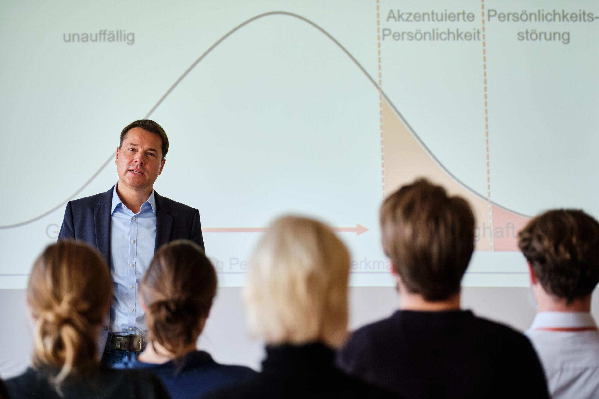 Dozent hält medizinischen Vortrag vor Teilnehmenden, Präsentationsfolie zeigt Kurve zu Persönlichkeitsmerkmalen