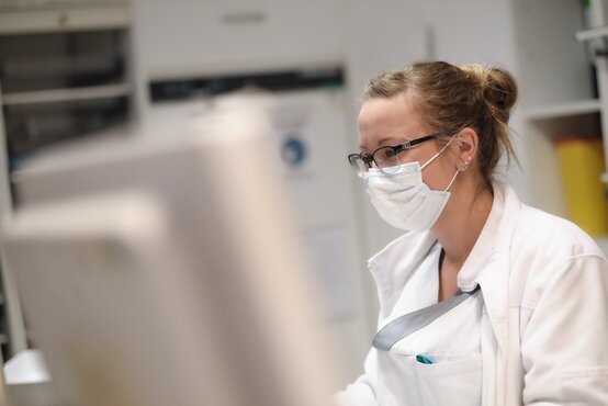 Junge Frau im weißen Kittel mit Schutzmaske arbeitet konzentriert am Computer in medizinischer Einrichtung