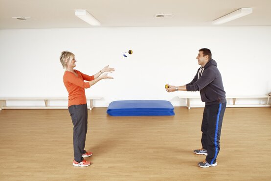 Frau und Mann jonglieren mit Bällen im hellen Sportraum der Klinik während einer Bewegungstherapie