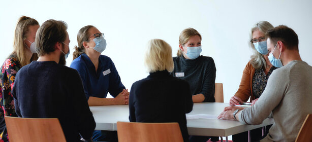 Teammeeting mit sieben Mitarbeitenden in heller Besprechung, alle mit medizinischen Masken und Abstand