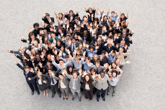 Große Gruppe von Menschen in Businesskleidung steht auf Kiesboden und winkt zur Kamera nach oben