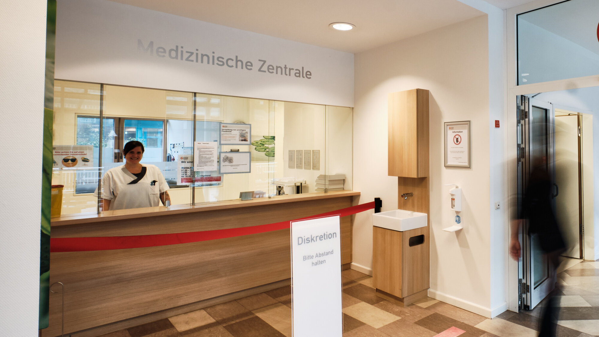 Lächelnde Mitarbeiterin im weißen Kittel hinter Glasscheibe an medizinischer Rezeption, Patient betritt Empfangsbereich