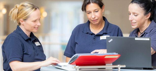 Drei Pflegekräfte im Team besprechen Patientendokumente am Schreibtisch mit Laptop