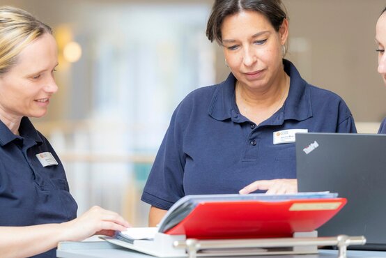Drei Pflegekräfte im Team besprechen Patientendokumente am Schreibtisch mit Laptop