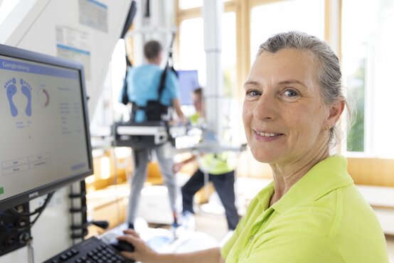 Älterer Mann trainiert mit robotischem Gangtrainer, Im Vordergrund Frau die am Computer die Analyse betreut