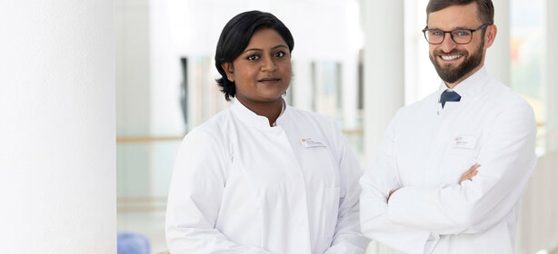 Zwei Ärzte in weißer Kleidung mit Namensschildern stehen freundlich in heller, moderner Klinik