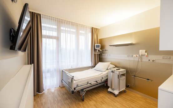 Modernes, helles Einzelzimmer mit elektrisch verstellbarem Krankenbett, Nachttisch, Flachbildfernseher und großen Fenstern