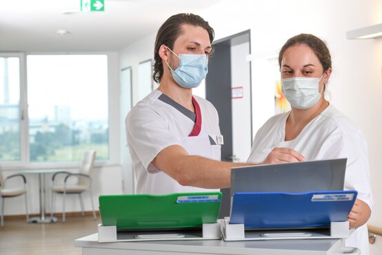 Zwei Pflegefachkräfte mit Schutzmasken im Gespräch an Computerarbeitsplatz