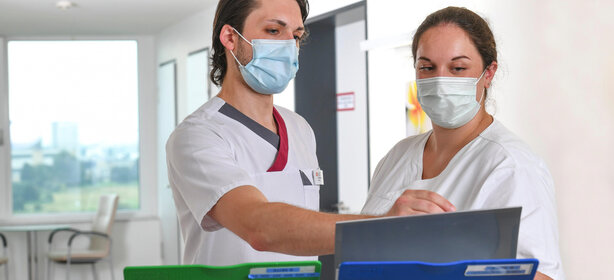 Zwei Pflegefachkräfte mit Schutzmasken im Gespräch an Computerarbeitsplatz